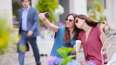 Açık hava kafesinde iki genç kız akıllı telefonlarıyla selfie çekiyor. Çantalarla alışveriş yaptıktan sonra açık kafede kahve içip akıllı telefon kullanan iki kadın.