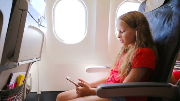 飛行機で旅行する愛らしい少女。子供は飛行機の窓の近くに座って音楽を聞く — ストック動画