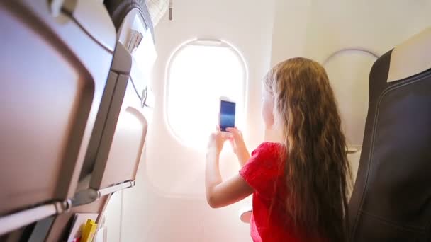 窓の近くに座っている飛行機で旅行する愛らしい少女。飛行機の窓の近くに座っている雲と空の写真を撮る子供 — ストック動画