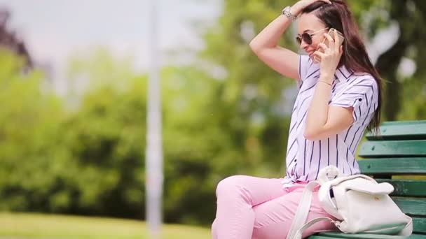 Joven chica caucásica hablando por su teléfono inteligente al aire libre. Hermosa chica en gafas de sol en la calle utilizando el teléfono inteligente y hablando por teléfono con su amigo — Vídeos de Stock