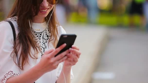 Jeune femme caucasienne envoyer un message par son smartphone en plein air dans le parc au coucher du soleil. Belle fille en lunettes de soleil assise sur un banc en bois à l'aide d'un smartphone — Video