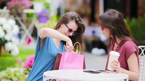Δύο νεαρά κορίτσια στο υπαίθριο καφέ. Δύο γυναίκες μετά τα ψώνια με τσάντες που κάθονται σε openair cafe με καφέ και τη χρήση smartphone — Αρχείο Βίντεο