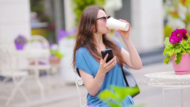 Retrato de mulher bonita sentada no café ao ar livre bebendo café e usando smartphone. Menina olhando instagram e beber delicioso cappuccino doce . — Vídeo de Stock