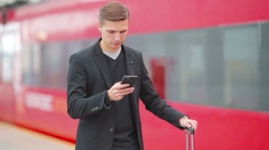 İstasyonda trenle seyahat eden akıllı telefon ve bavullu beyaz bir genç.