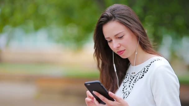 Mulher caucasiana jovem enviando mensagem por seu smartphone ao ar livre no parque. Menina bonita em óculos de sol sentado em banco de madeira usando smartphone — Vídeo de Stock