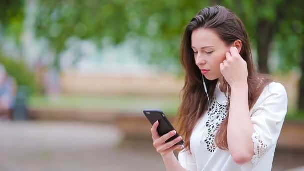 Ung kaukasisk kvinna skickar meddelande via sin smartphone utomhus i parken. Vacker flicka i solglasögon sitter på träbänk med hjälp av smartphone — Stockvideo