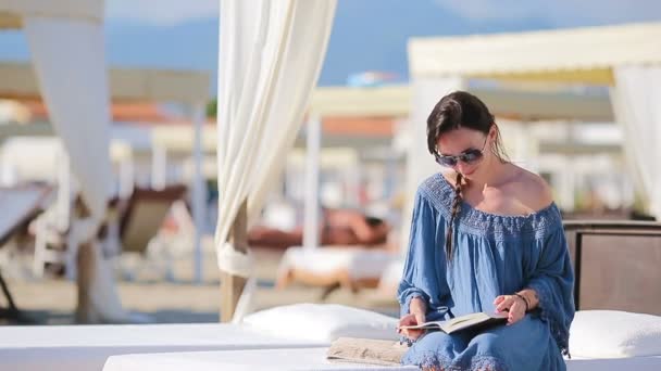 Молодая женщина читает книгу во время тропических пляжных каникул. Девушка из моды читает сидя на белых шезлонгах на европейском пляже — стоковое видео