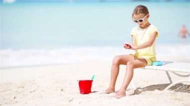 Tropikal plajda oturan güneş kremi şişeli küçük kız.
