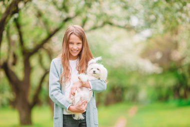 Küçük gülümseyen kız parkta oynaşıyor ve yavru köpeğe sarılıyor.