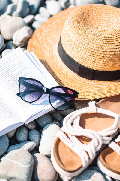 Chapéu de praia em livro aberto com protetor solar e sapatos na praia de seixos — Fotografia de Stock