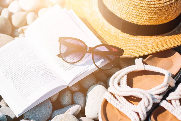 Sombrero de playa en libro abierto con protector solar y zapatos en la playa de guijarros —  Fotos de Stock