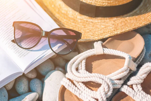 Sombrero de playa en libro abierto con protector solar y zapatos en la playa de guijarros —  Fotos de Stock