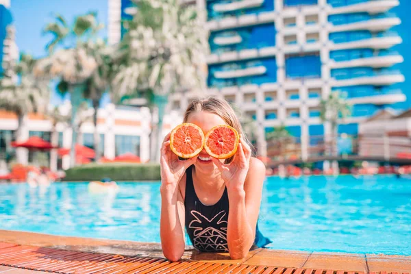 Niña cubriendo los ojos con mitades naranjas cerca de los ojos en la piscina de fondo — Foto de Stock