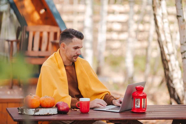 Šťastný mladý muž pracující na notebooku a pití kávy sedí u dřevěného stolu venku — Stock fotografie