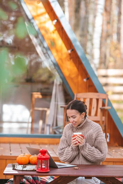 Szczęśliwa młoda kobieta pracuje na laptopie i pije kawę siedząc przy drewnianym stole na świeżym powietrzu — Zdjęcie stockowe