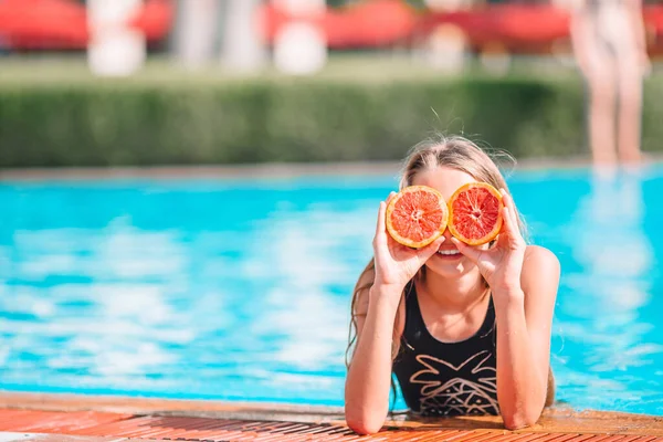 Niña cubriendo los ojos con mitades naranjas cerca de los ojos en la piscina de fondo — Foto de Stock