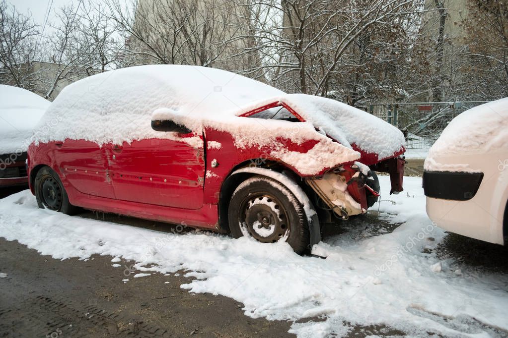 Damaged red car, with a bent hood, covered with snow, sitting in an auto service yard, after being involved in a road accident