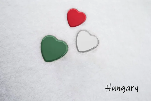 Cores da bandeira húngara (Framboesa Francesa, Branco, Amazônia / verde) pintadas em três corações. Fundo de neve com o país, Hungria, escrito no canto inferior direito. Conceito para boas-vindas calorosas no turismo . — Fotografia de Stock