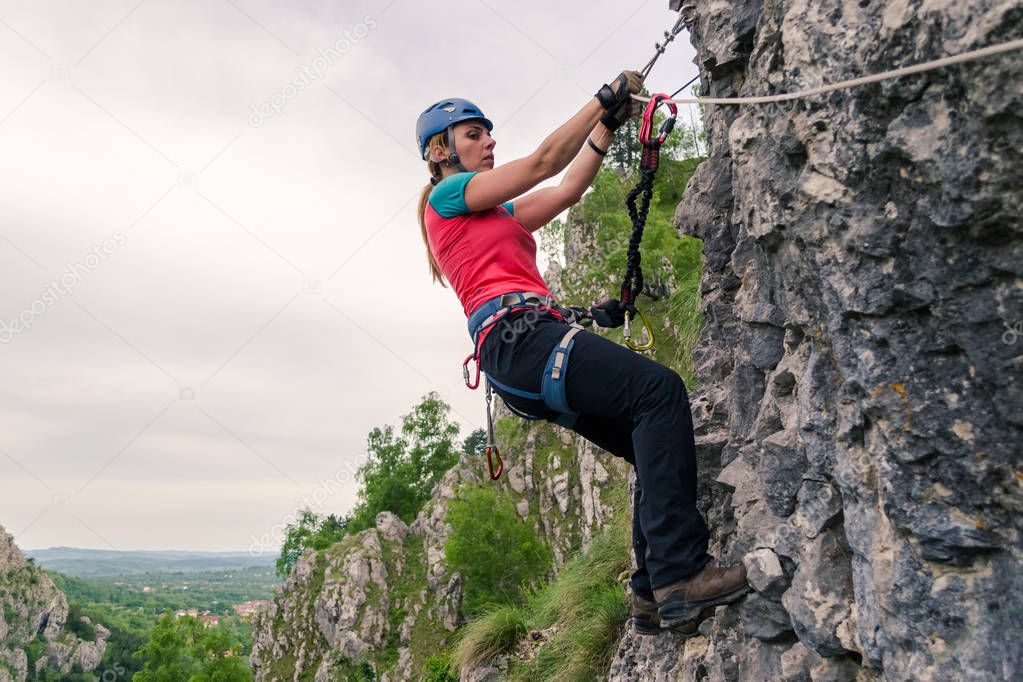 Young woman hanging from a via ferrata cable, while passing a difficult section, equipped with helmet, harness, kit. Klettersteig route at Baia de Fier, Pestera Muierilor, Romania, Gorj county.