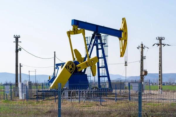 Blue and yellow pump jack above an oil well surrounded by electricity poles and protected with a fence, in bright daylight, in Summer, near Mol gas station - Ploiesti city, Romania.