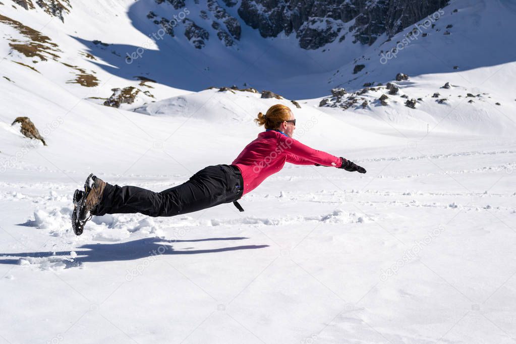 Joyful female hiker jumping on the snow, horizontally, resembling a Superman/Superwoman levitating pose, near Malaiesti hut, in Bucegi/Carpathian mountains, Romania, on a bright late Winter day.