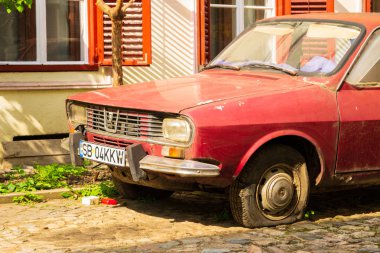 Eski, paslı, kırmızı Dacia 1300 otomobil, düz bir lastik ama aktif plakalı, parke taşı ile bir avluda güneşte park edilmiş. Kısmi ön ve yan görünüm.