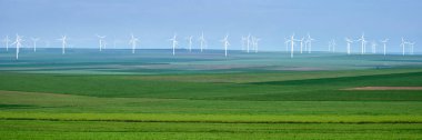 Romanya'nın Dobrogea kentinde, bulutlu bir gökyüzüüzerinde, çeşitli yeşil tonlarında tarım ürünlerinin katmanlarında beyaz rüzgar türbinlerinin panoraması. Sürdürülebilir, yeşil, enerji üretimi kavramı.