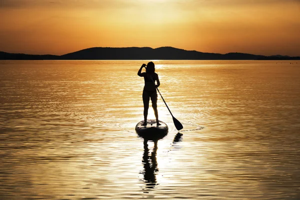 Hırvatistan, Adriyatik Denizi, Sibenik yakınlarındaki bir stand up paddleboard (Sup) gün batımında kürek çeken kadın silueti. Turizm konsepti.