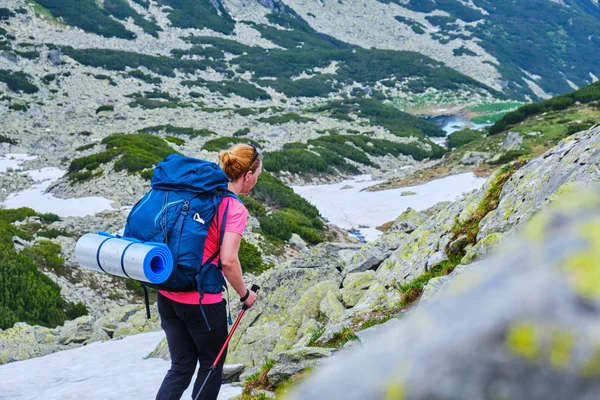 Woman with heavy backpack and a rolled sleeping pad, navigating a trekking trail in Retezat mountains (part of Carpathians), Romania, during Summer hiking season, with patches of snow still present.