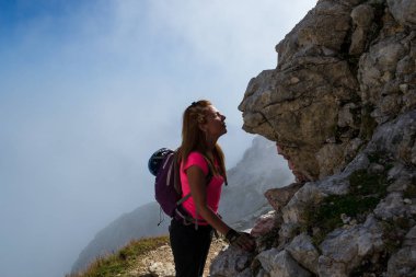 Woman hiker kissing a rock face in the Julian Alps, Slovenia, just under Mangart peak, after descending the via ferrata route. Misty clouds in the back. clipart