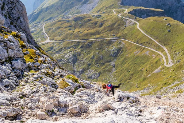 Ontspannen vrouw poseren grappig met haar been omhoog, hoog op een bergpad boven Mangart zadel, met Mangart weg zichtbaar ver onder, op een heldere dag, tijdens een via ferrata/wandeltocht. — Stockfoto