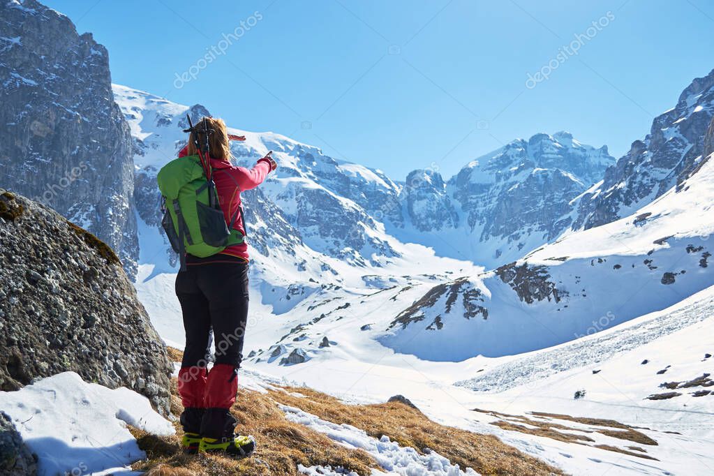 Woman hiker pointing towards Malaiesti Valley and Hornul Mare chimney, in Bucegi mountains, Romania, on a sunny Winter day, with a small avalanche on a slope nearby, on the right.