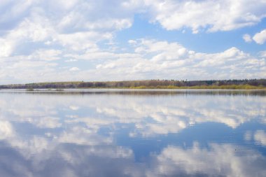Mavi gökyüzü ve bulutlar nehre yansıdı. Su yüzeyinde dalgalanmalar. Ufukta orman
