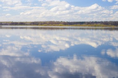 Mavi gökyüzü ve bulutlar nehre yansıdı. Su yüzeyinde dalgalanmalar. Ufukta orman