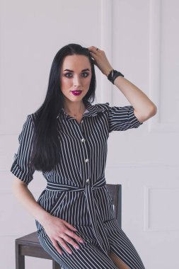 Çizgili bir elbise seksi esmer model yüksek bir sandalyede oturuyor. Kız saçını düzeltiyor. Arka planda beyaz duvar.