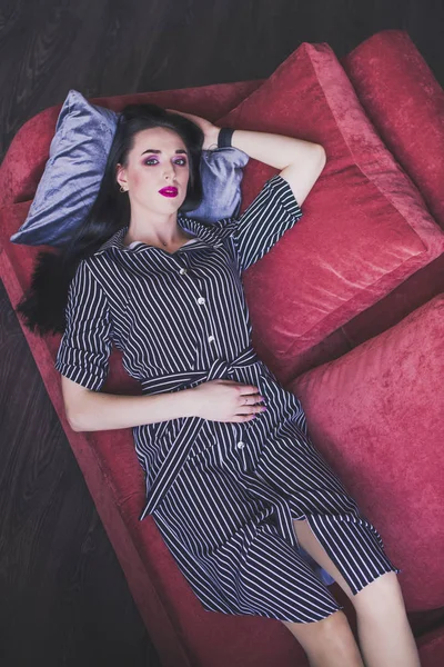 Brunetka model w paski sukienka leżąca na czerwonej kanapie — Zdjęcie stockowe