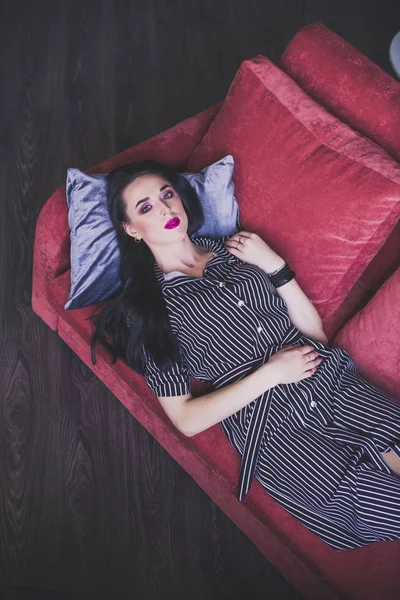 Brunetka model w paski sukienka leżąca na czerwonej kanapie — Zdjęcie stockowe