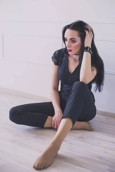Modelo morena descalça em um terno preto sentado no chão. De mãos dadas. Contra o fundo de uma parede branca . — Fotografia de Stock
