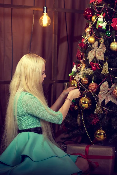 Modelo loira bonita em um vestido turquesa veste-se uma árvore de Natal. Decoração de casa quente. Atmosfera de Natal. Tonificação retrô . — Fotografia de Stock