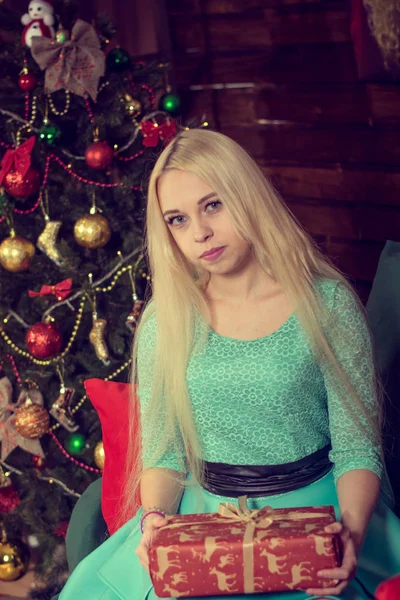 Das hübsche blonde Model in einem türkisfarbenen Kleid öffnet ein Weihnachtsgeschenk. warme Wohnkultur. Weihnachtsstimmung. Geschenke für Weihnachten und Neujahr. — Stockfoto