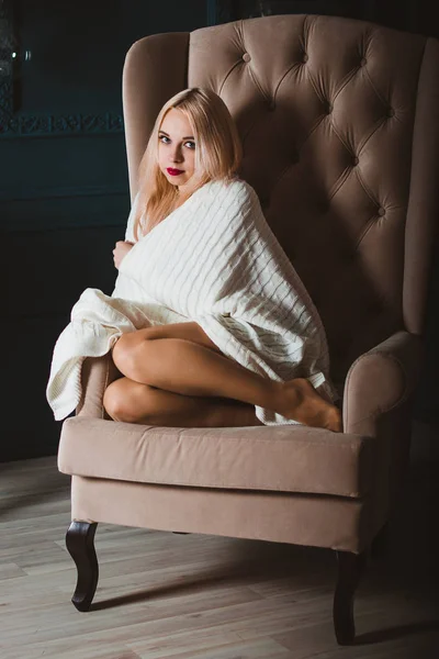 Sexy blondine model zit in een stoel met hoge rug, bedekt met een Plaid. Mooie poten zijn blootgesteld aan onder de Plaid. Klassiek interieur in de rechter. — Stockfoto