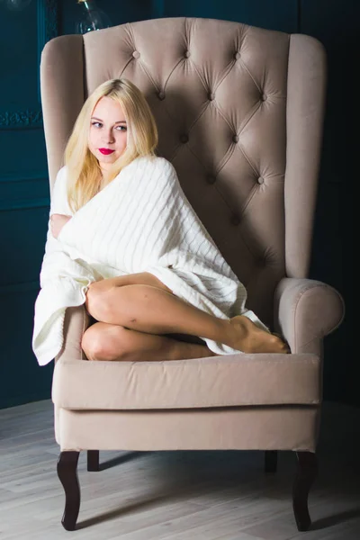 Sexy blondine model zit in een stoel met hoge rug, bedekt met een Plaid. Mooie poten zijn blootgesteld aan onder de Plaid. Klassiek interieur in de rechter. — Stockfoto