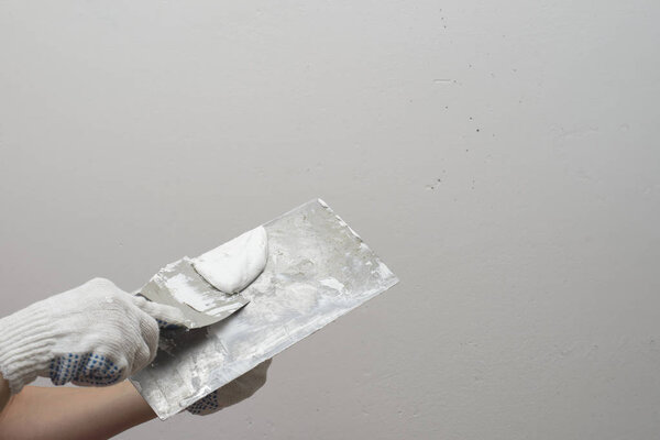 A hand with a small spatula smears the putty on a large spatula. Against the background of white plastered wall