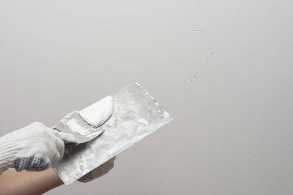 A hand with a small spatula smears the putty on a large spatula. Against the background of white plastered wall