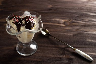 Beyaz vanilyalı dondurma topları tatlı bardağına çikolata soslu ve kaşıklı. Kaba ahşaptan yapılmış eski bir masada süslenmiş dondurma. Kopyalama alanı.