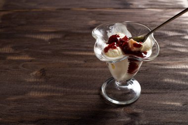 Beyaz vanilyalı dondurma topları ile ahududu reçeli tatlı bardağında kaşık. Kaba ahşaptan yapılmış eski bir masada süslenmiş dondurma. Kopyala-uzayı düzlemi.