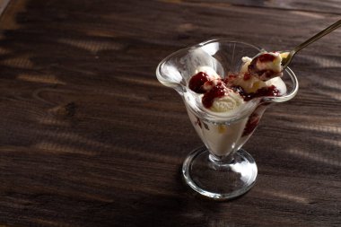 Beyaz vanilyalı dondurma topları ile ahududu reçeli tatlı bardağında kaşık. Kaba ahşaptan yapılmış eski bir masada süslenmiş dondurma. Kopyala-uzayı düzlemi.