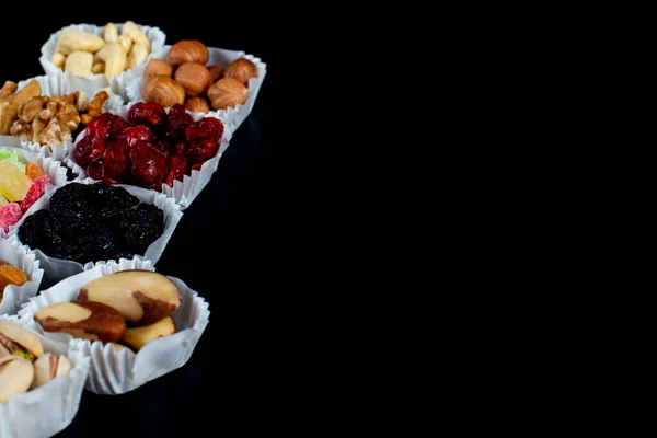 A serving of assorted candied fruit, dried cherries, almonds, raisins, walnuts and hazelnuts in paper muffin cups on a black background. Copyspace.