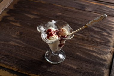Beyaz vanilyalı dondurma topları ile ahududu reçeli tatlı bardağında kaşık. Kaba ahşaptan yapılmış eski bir masada süslenmiş dondurma. Kopyala-uzayı düzlemi.