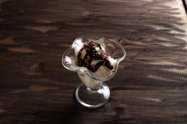 Beyaz vanilyalı dondurma topları tatlı bardağına çikolata soslu ve kaşıklı. Kaba ahşaptan yapılmış eski bir masada süslenmiş dondurma. Kopyalama alanı.